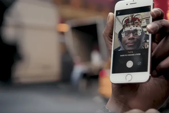 A hand holding up an iPhone on a New York street, the HamCam crown filter on the fan’s face